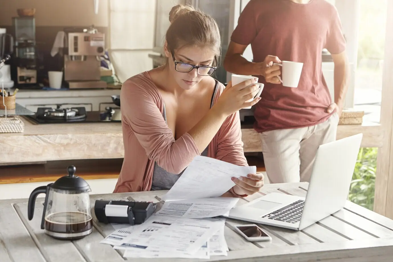 Eine junge Familie hat mit finanziellen Problemen zu kämpfen. Die ernste Ehefrau mit Brille sitzt vor einem Laptop, geht Rechnungen durch und hält eine Tasse.